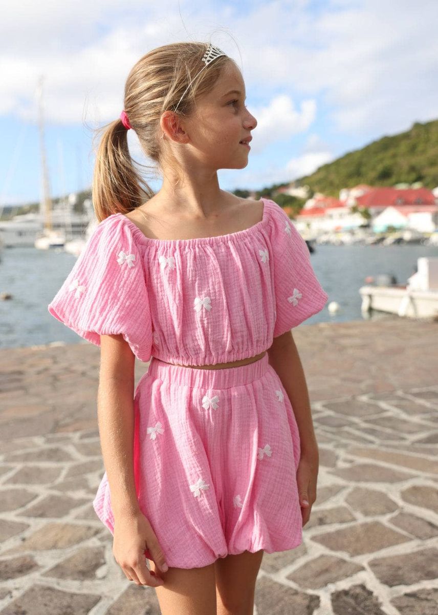 Young girl in a pink lola and the boys outfit standing by a waterfront with boats and buildings in the background.