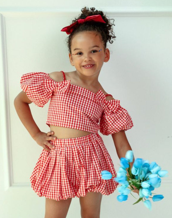 Young girl in a lola and the boys red checkered outfit holding blue flowers against a white background