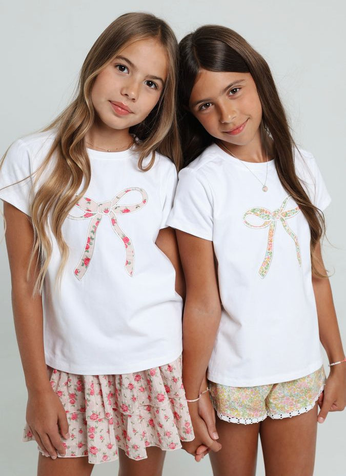 Two young girls wearing matching white t-shirts with floral designs and shorts on a plain background
