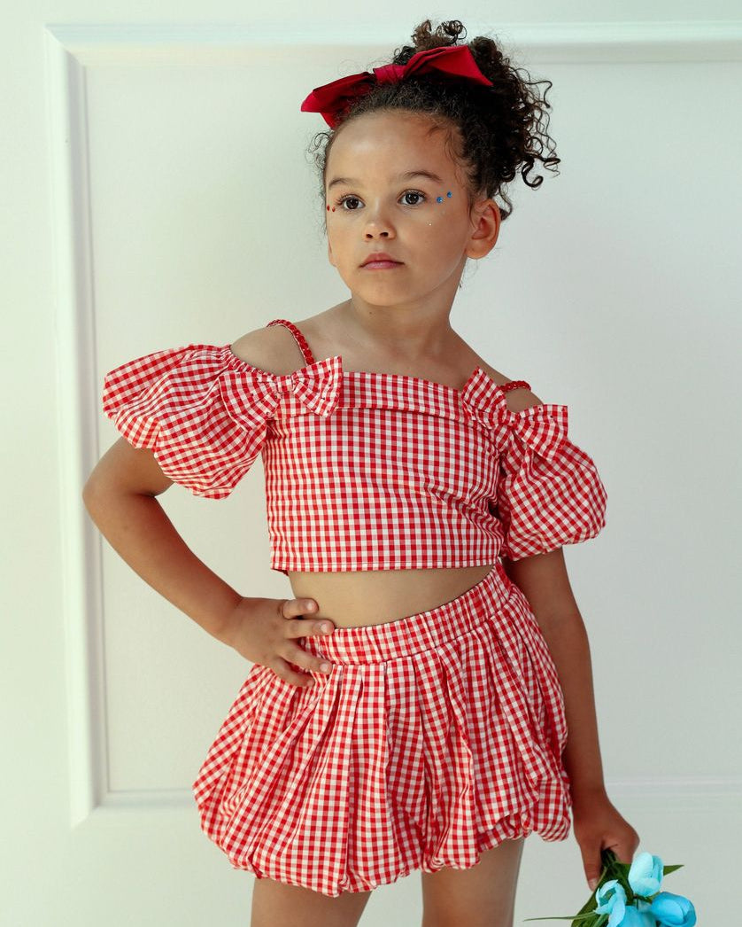 Young girl wearing a red and white checkered outfit with a bow in her hair, standing against a white background.