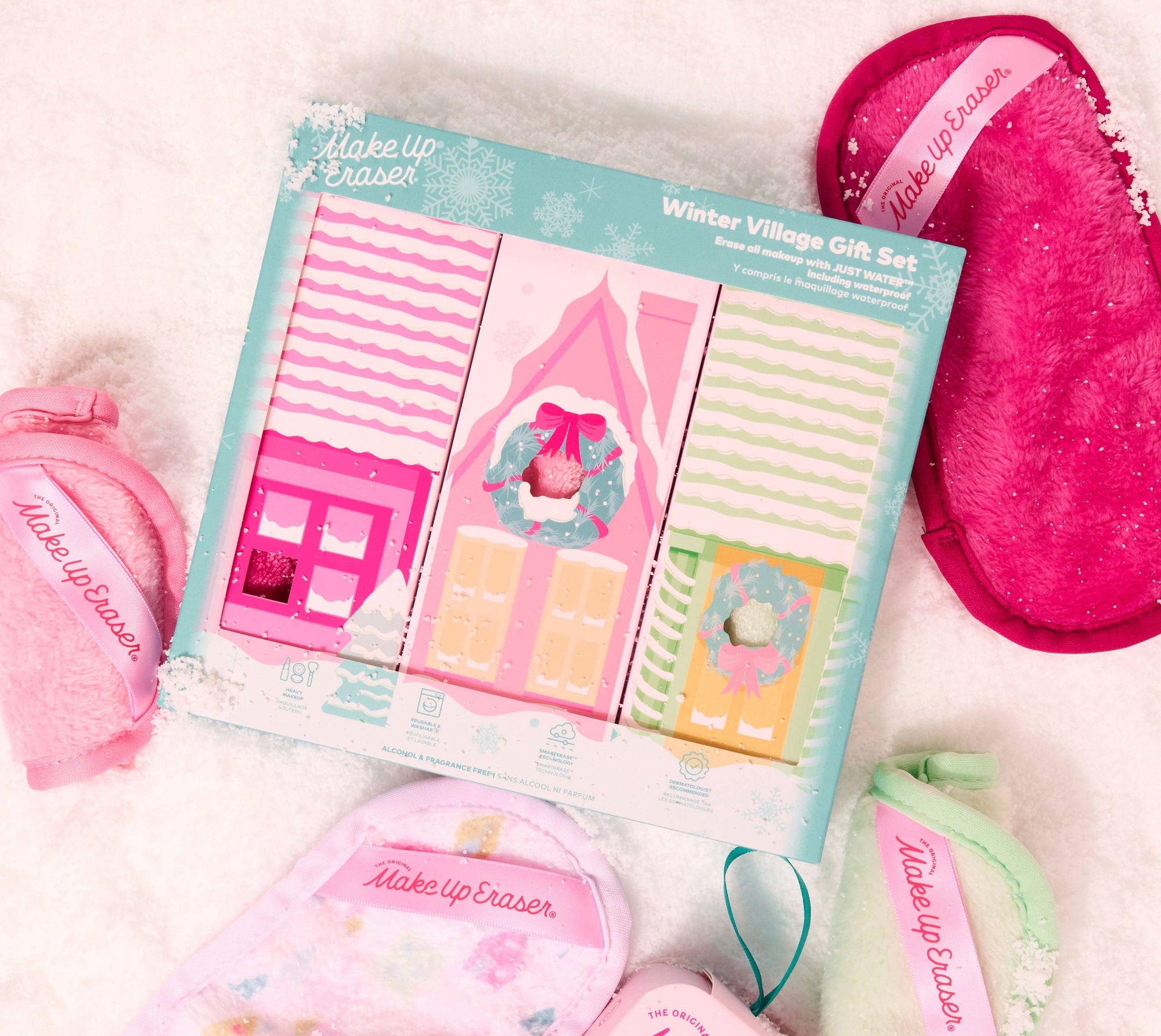 A Make Up Easel Winter Village Gift Set arranged on a white textured surface. The set includes a pink plush bath mat, a pink plush towel, a pink plush mitt, a mint green plush mitt, a pink dollhouse-shaped soap dish, striped bath towels, and a packaged set of decorative bath blocks inside a light blue box.