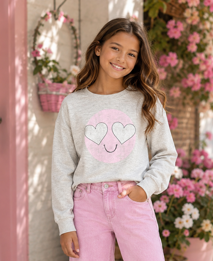 A young girl stands on a sunlit sidewalk wearing a light gray sweatshirt with a pink heart graphic, pink wide-leg jeans, and white sneakers. Pink flowers and a pink door are visible in the background.