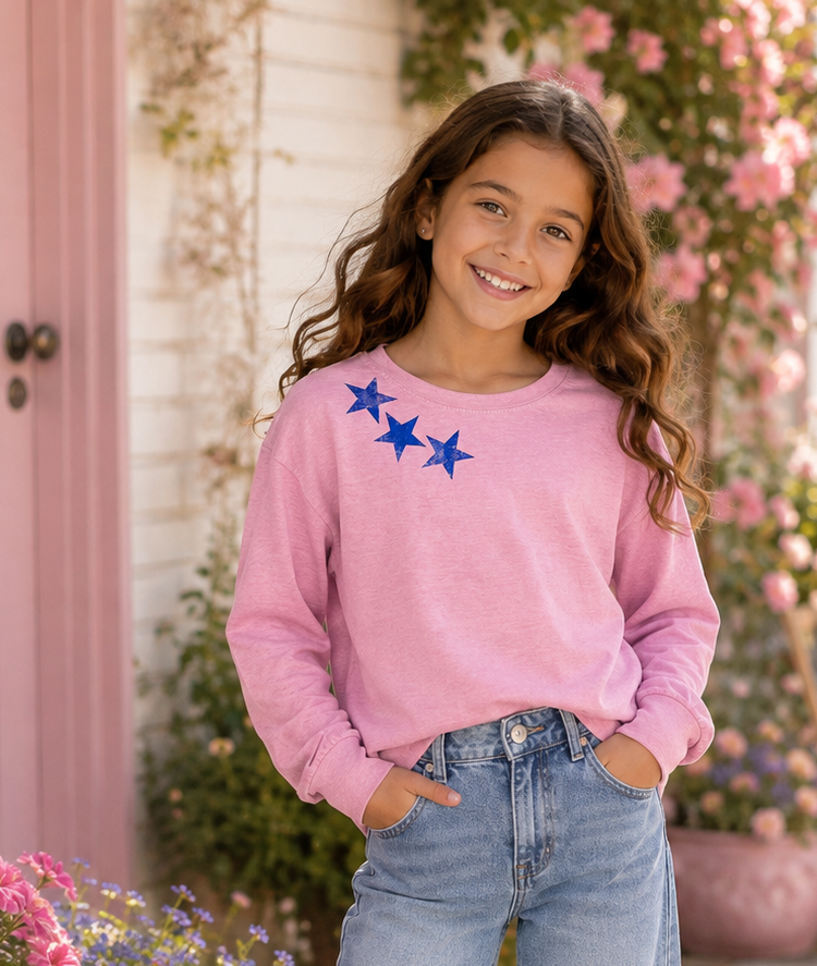 A young girl stands on a sunlit porch wearing a pink long-sleeve sweatshirt with three blue stars on the chest, light-wash wide-leg jeans, and white sneakers. Pink flowers and a pink door are visible in the background.