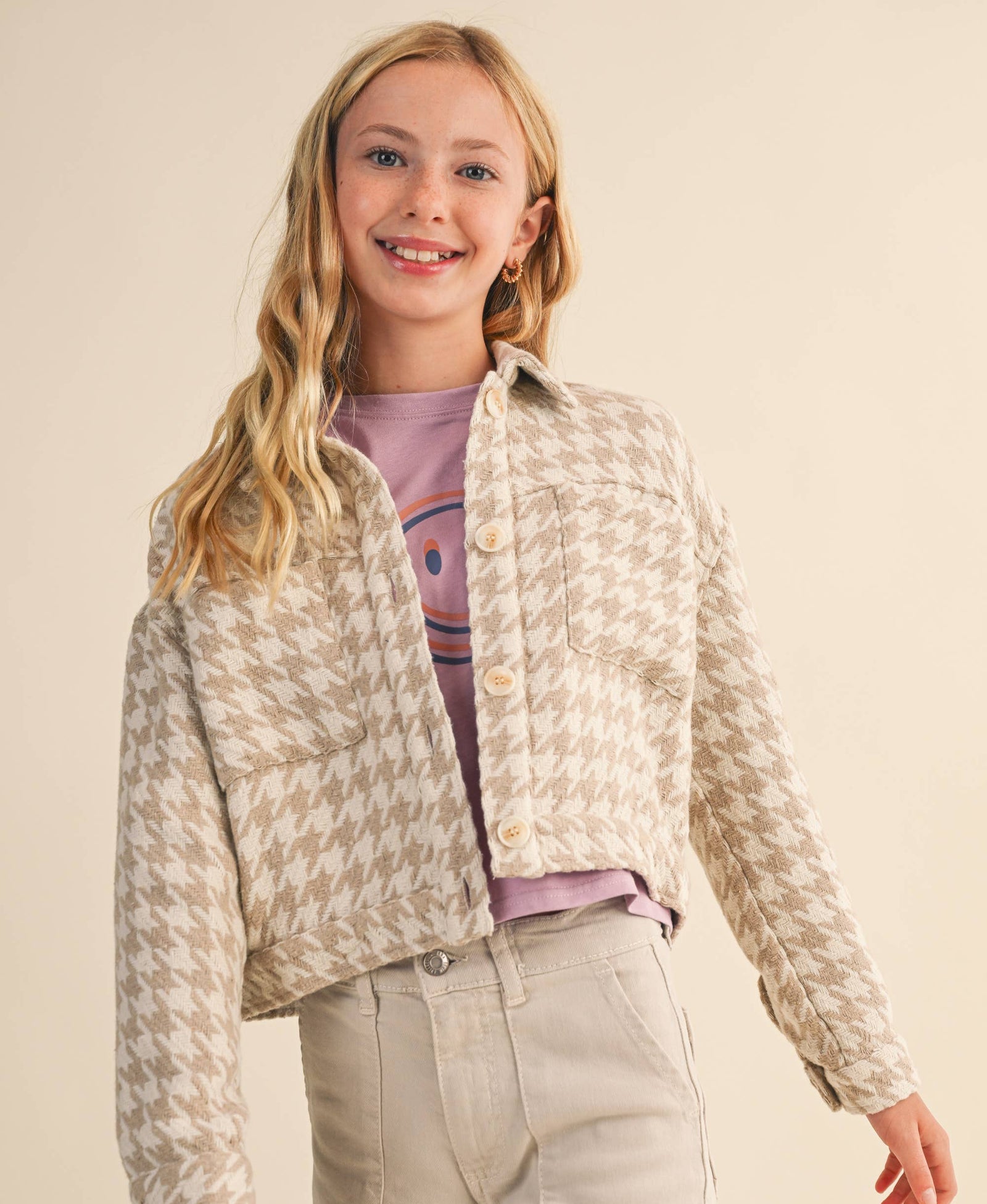 A young girl with blonde hair wearing a beige and white houndstooth cropped jacket over a purple t-shirt and light beige pants, standing against a solid beige background.