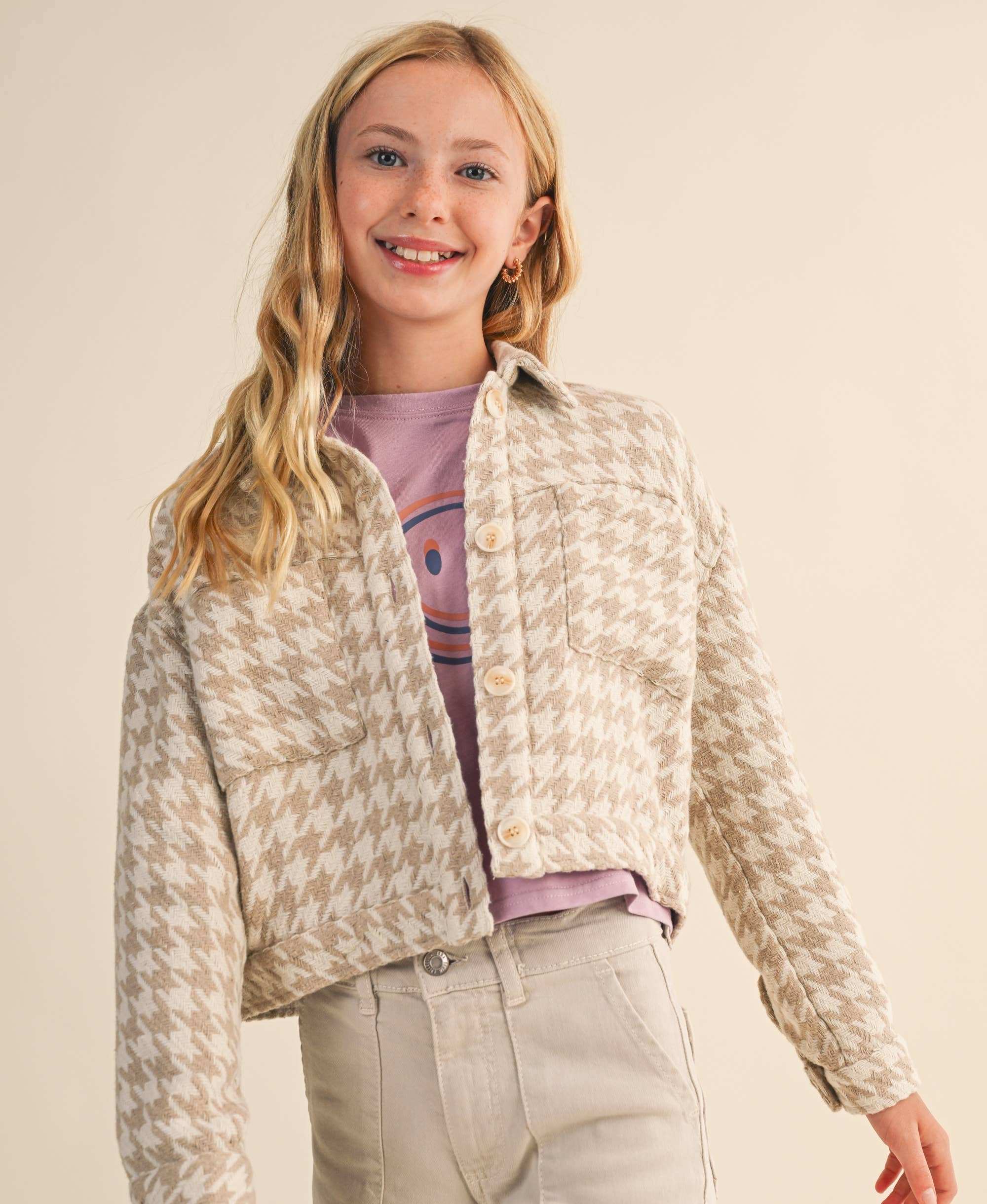 A young girl with blonde hair wearing a beige and white houndstooth cropped jacket over a purple t-shirt and light beige pants, standing against a solid beige background.