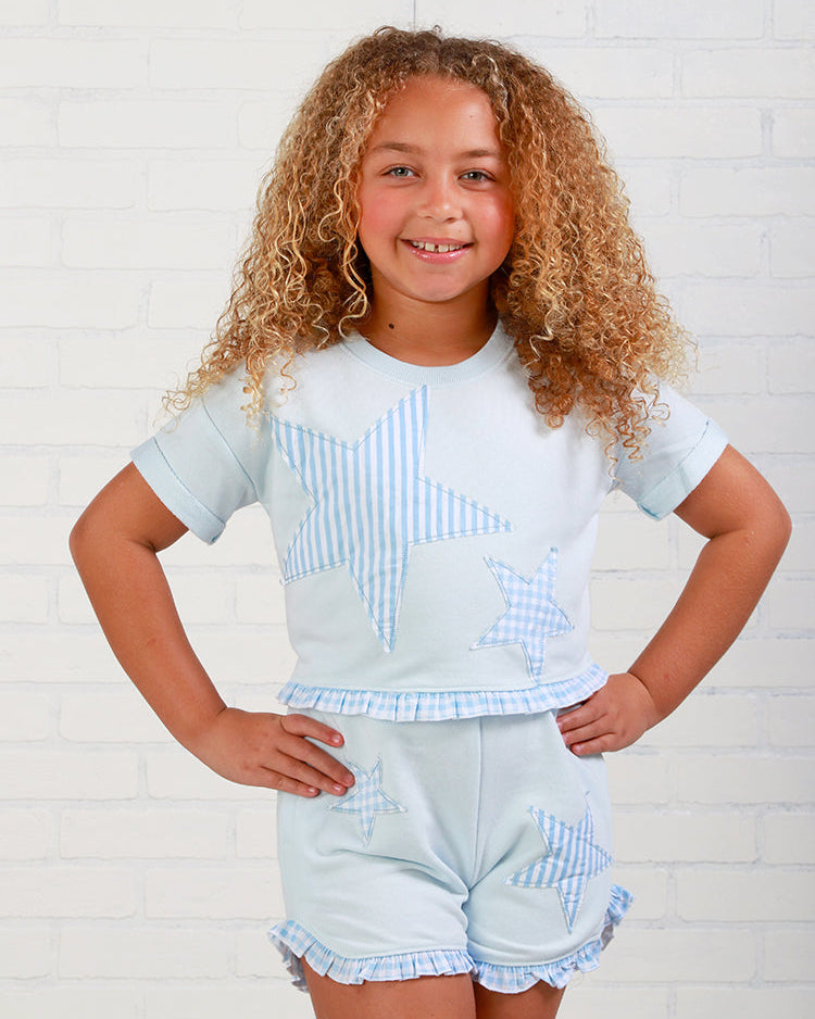Young girl wearing a light blue star-patterned shirt and shorts against a white brick wall.