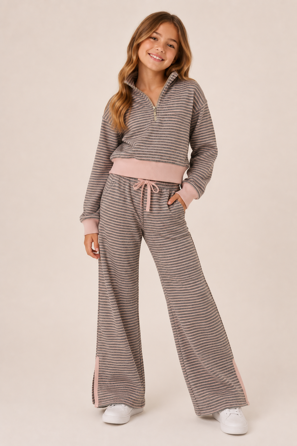 A young girl modeling a matching two-piece loungewear set consisting of a gray and white striped half-zip pullover and wide-leg pants with a light pink waistband, drawstring, and side panels, paired with white sneakers against a solid beige background.