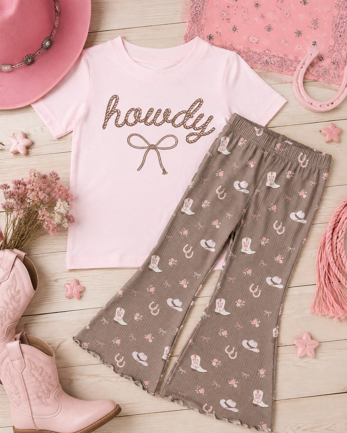 A toddler girl's western-themed two-piece outfit featuring a light pink short-sleeve t-shirt with 'howdy' and a bow in brown glitter, paired with brown flared leggings printed with pink and white cowboy boots, hats, and horseshoes. The outfit is styled in a flat lay with pink cowboy boots, a pink hat, lace, and wooden stars on a light wood background.