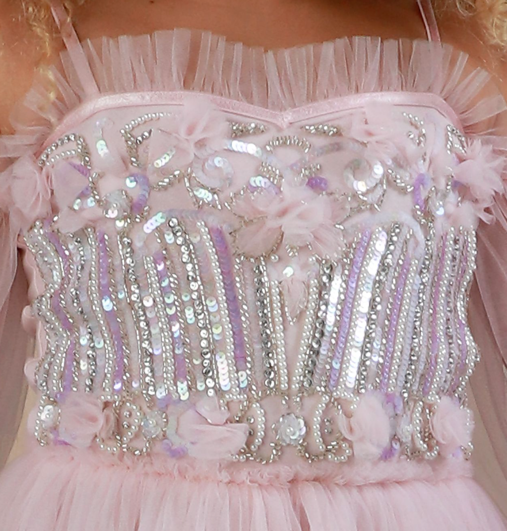A close-up of a light pink embellished dress bodice  by Oohlala Couture with tulle ruffles, sequins, beads, and floral appliqué. The front is covered in silver and iridescent sequins with pink and lavender floral accents.