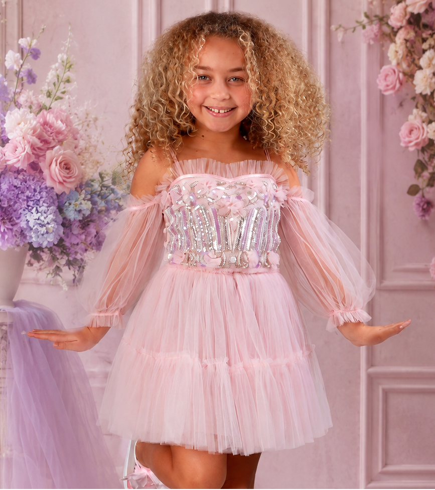 A young girl with curly blonde hair wearing a light pink off-the-shoulder pink tulle party dress with a sequined bodice, posing in a studio setting with a decorative tree in a pot and a white brick wall background.