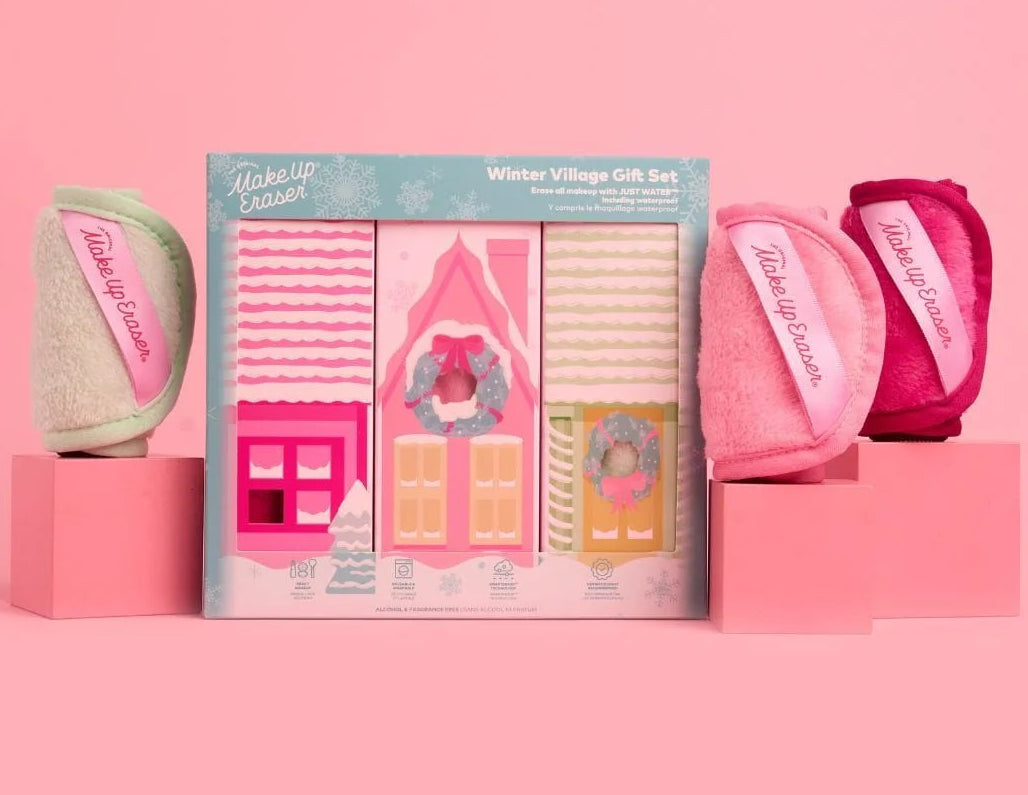 A boxed gift set displayed on a pink studio background, featuring a central illustrated package labeled 'Winter Village Gift Set' with three small bath towels or washcloths in pink, beige, and magenta arranged around it on pink blocks.