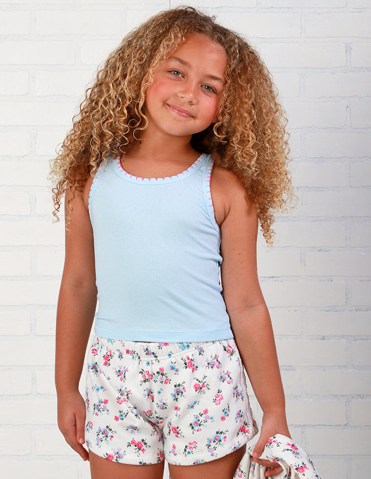 Young girl wearing a light blue  flowers by zoe tank top and floral shorts against a white brick wall.