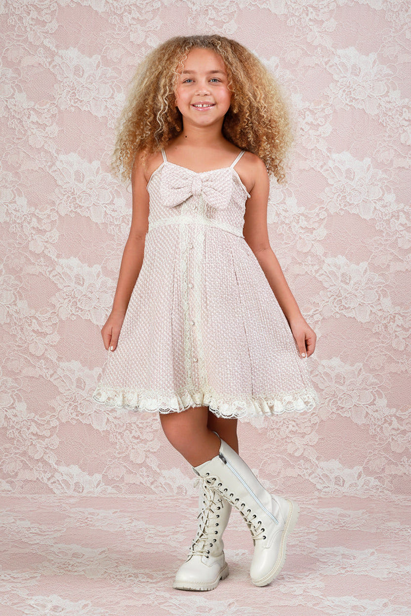A young girl with curly blonde hair stands wearing a cream-colored spaghetti strap dress with a large bow at the bust, a polka-dot pattern, and a ruffled hem. She is also wearing tall cream lace-up boots, posed in front of a white and light wood paneled wall.