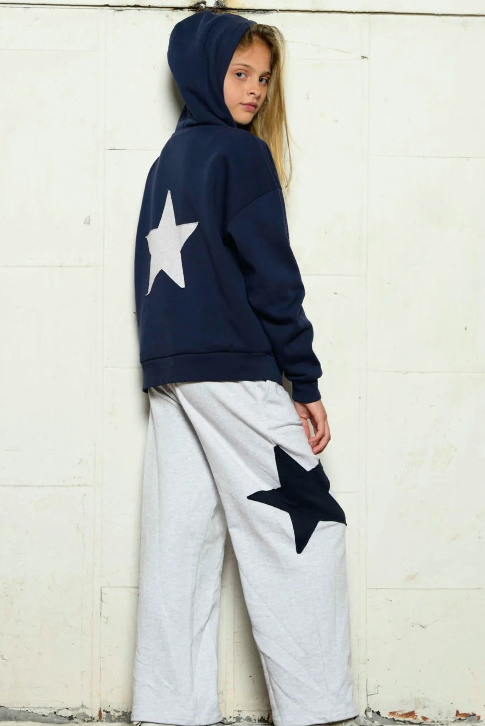 A child model wearing a navy blue hoodie by katiej nyc with a large white star on the back, paired with light gray wide-leg sweatpants featuring a large black star on the thigh, leaning against a white wooden wall.