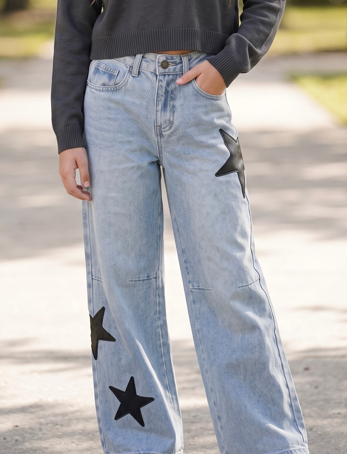 tween girtl wearing light-wash wide-leg jeans with black star patches on the legs by KatieJ NYC, paired with a dark gray long-sleeve top, standing outdoors on a paved path.