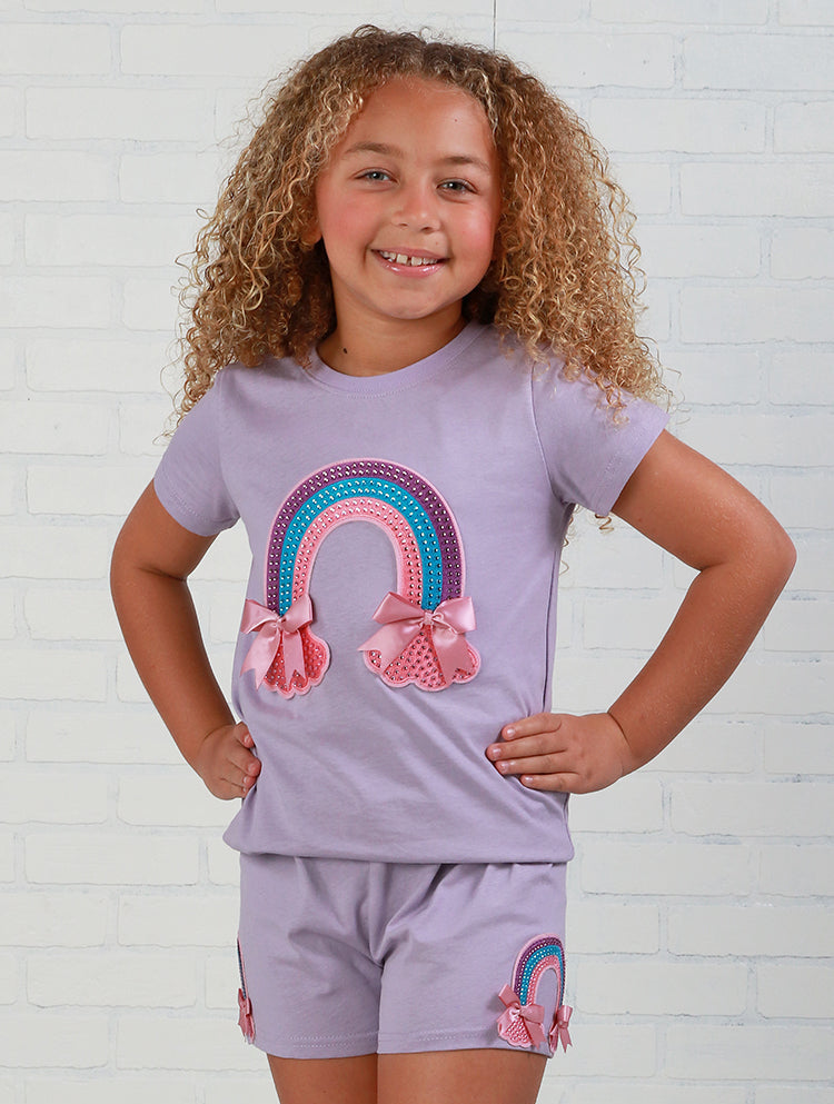 Young girl wearing a matching lavender outfit with rainbow and bow designs on a white brick wall background.