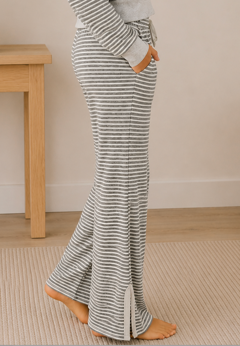 A person wearing gray and white striped lounge pants with a drawstring waist and side pockets, standing barefoot indoors on a textured rug near a light wood table.
