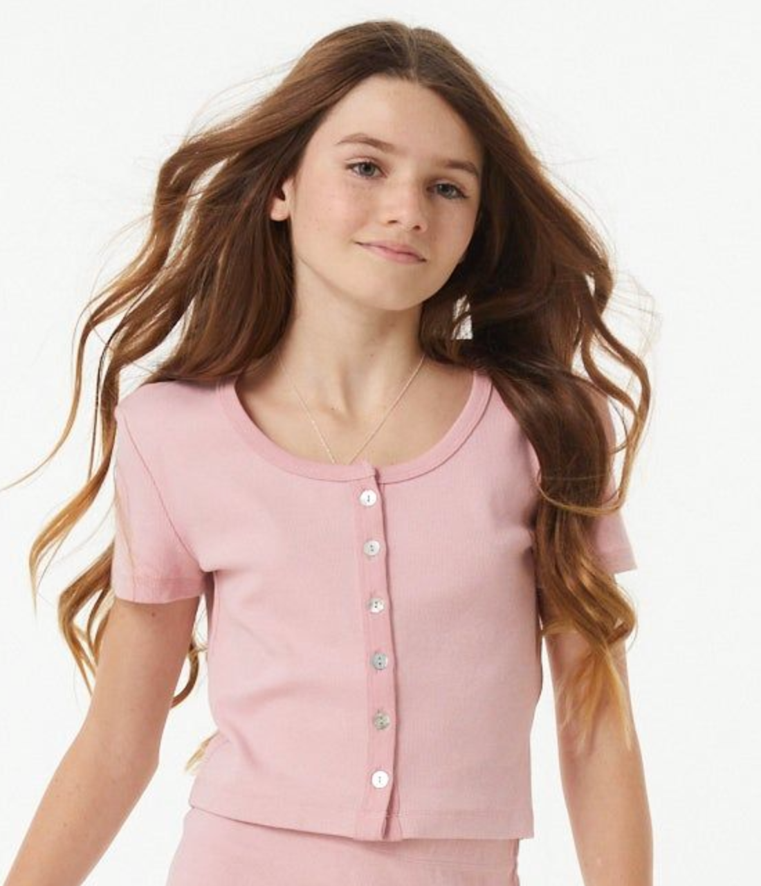A young girl with long wavy brown hair wearing a light pink short-sleeve button-up top and matching skirt, photographed against a clean white background.