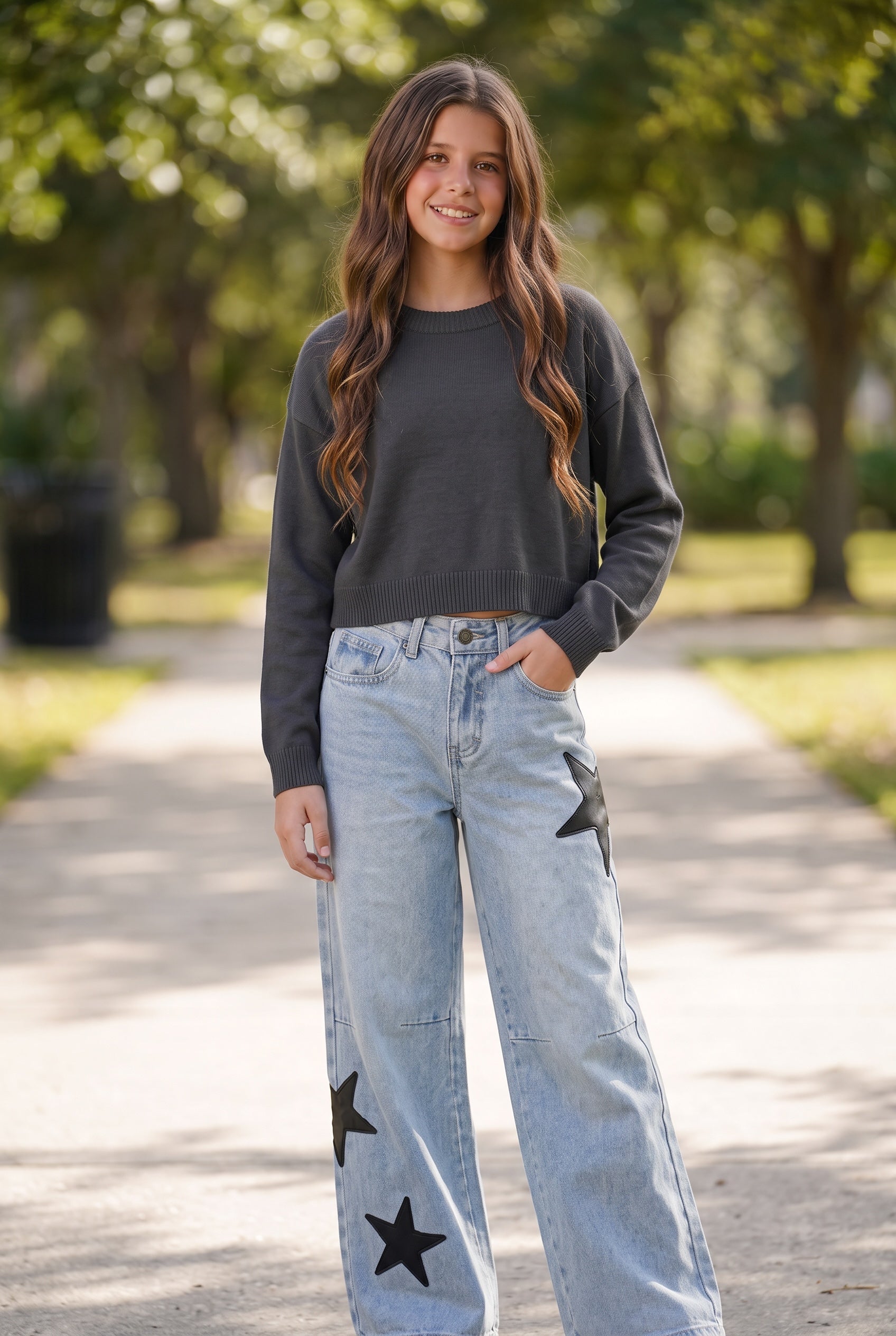 A tween girl stands on a sunlit park path wearing a dark charcoal gray long-sleeve crewneck sweatshirt and light-wash blue wide-leg jeans with black star patches by tween designer KatieJ.