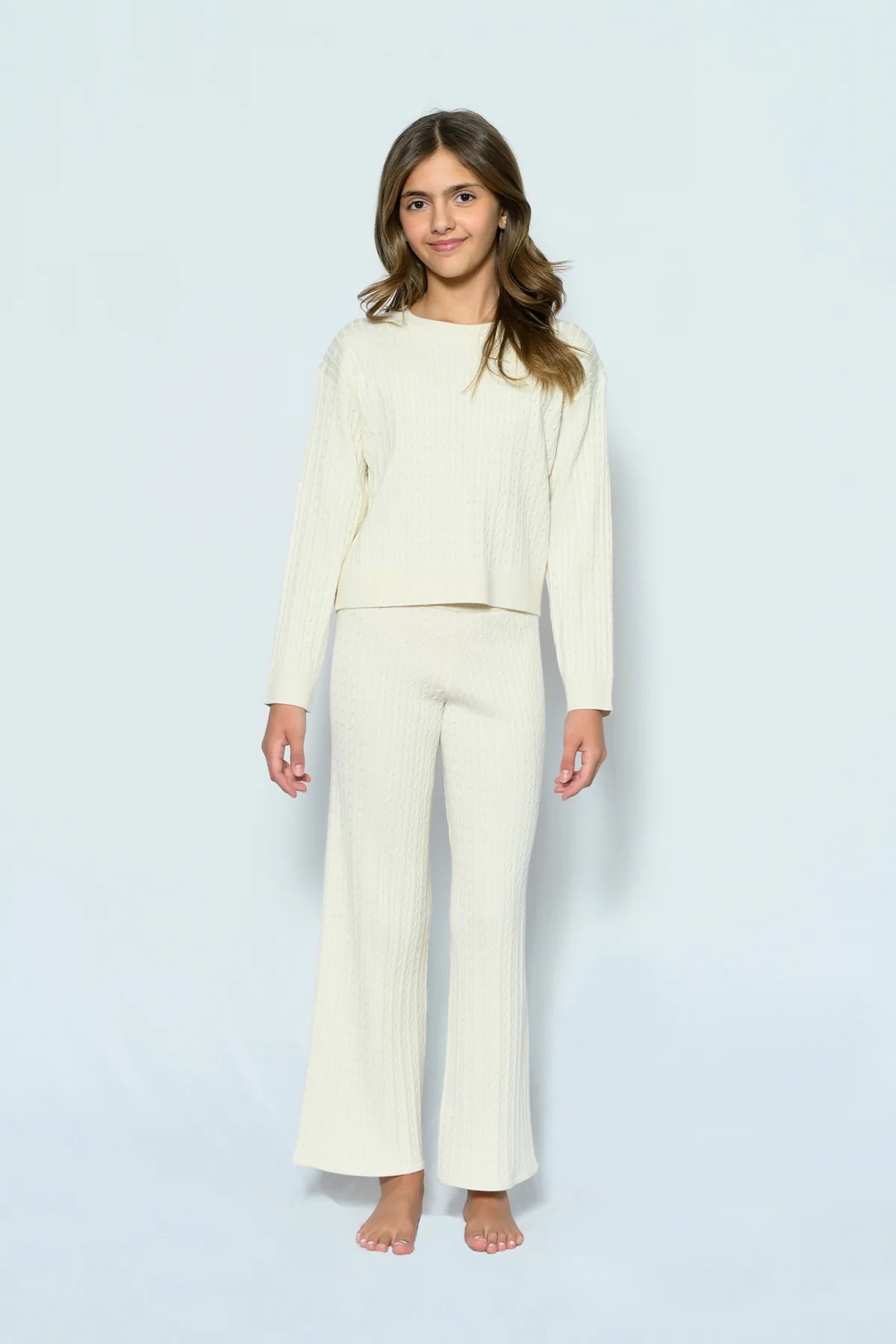 A young girl stands barefoot against a light blue studio background, wearing a matching cream-colored ribbed knit two-piece set consisting of a long-sleeve cropped top and wide-leg pants.