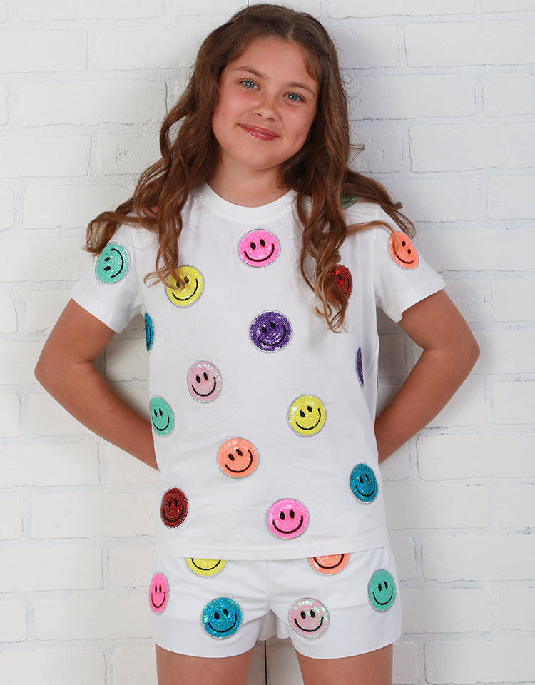 Young girl wearing a white outfit with colorful smiley face buttons against a white brick wall.