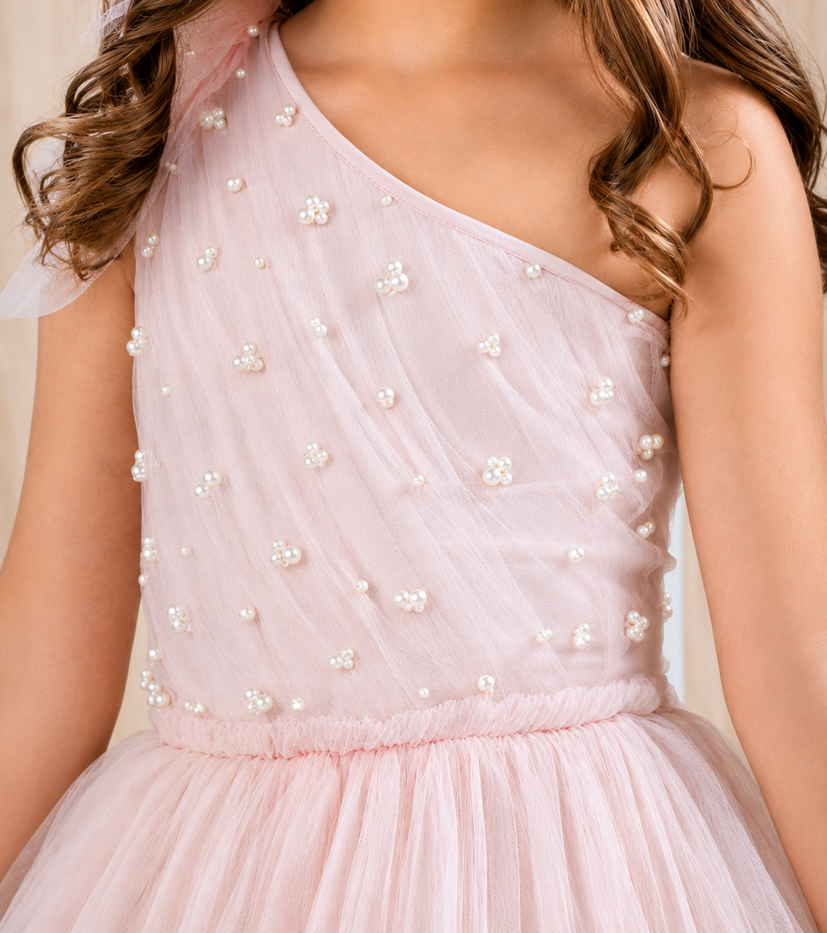 A close-up of a child wearing a light pink one-shoulder tulle dress with white pearl bead embellishments. The dress features a sheer upper bodice and a layered tulle skirt.