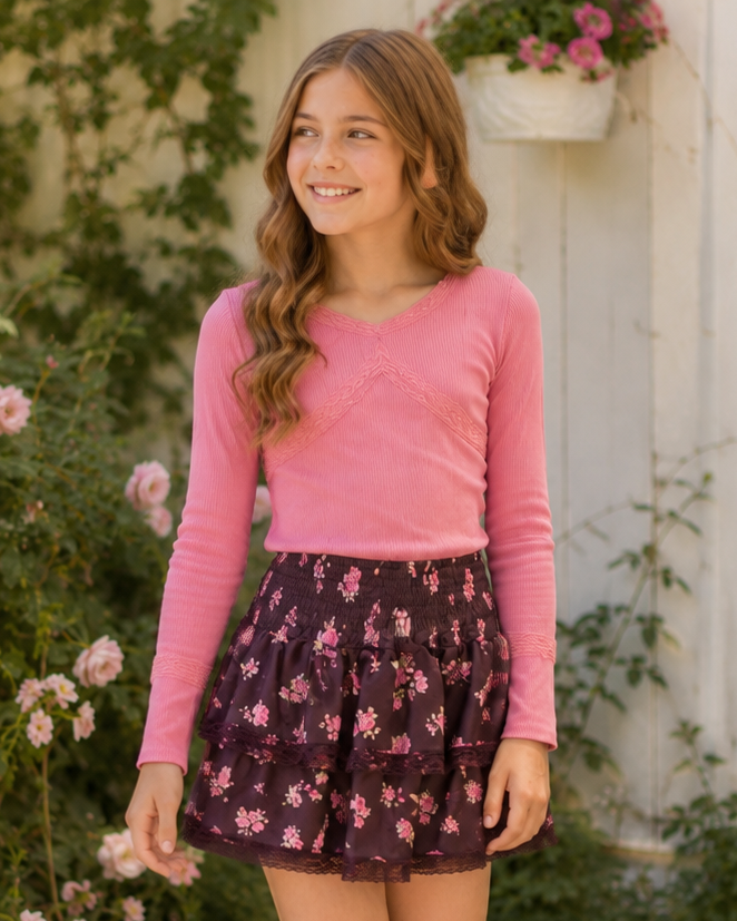 A young girl stands on a stone path in a garden setting, wearing a pink long-sleeve top and a dark purple floral mini skirt with white sneakers.