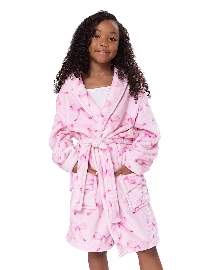 A young girl with curly dark hair wearing a pink plush bathrobe with a darker pink flamingo pattern, standing against a solid white background.