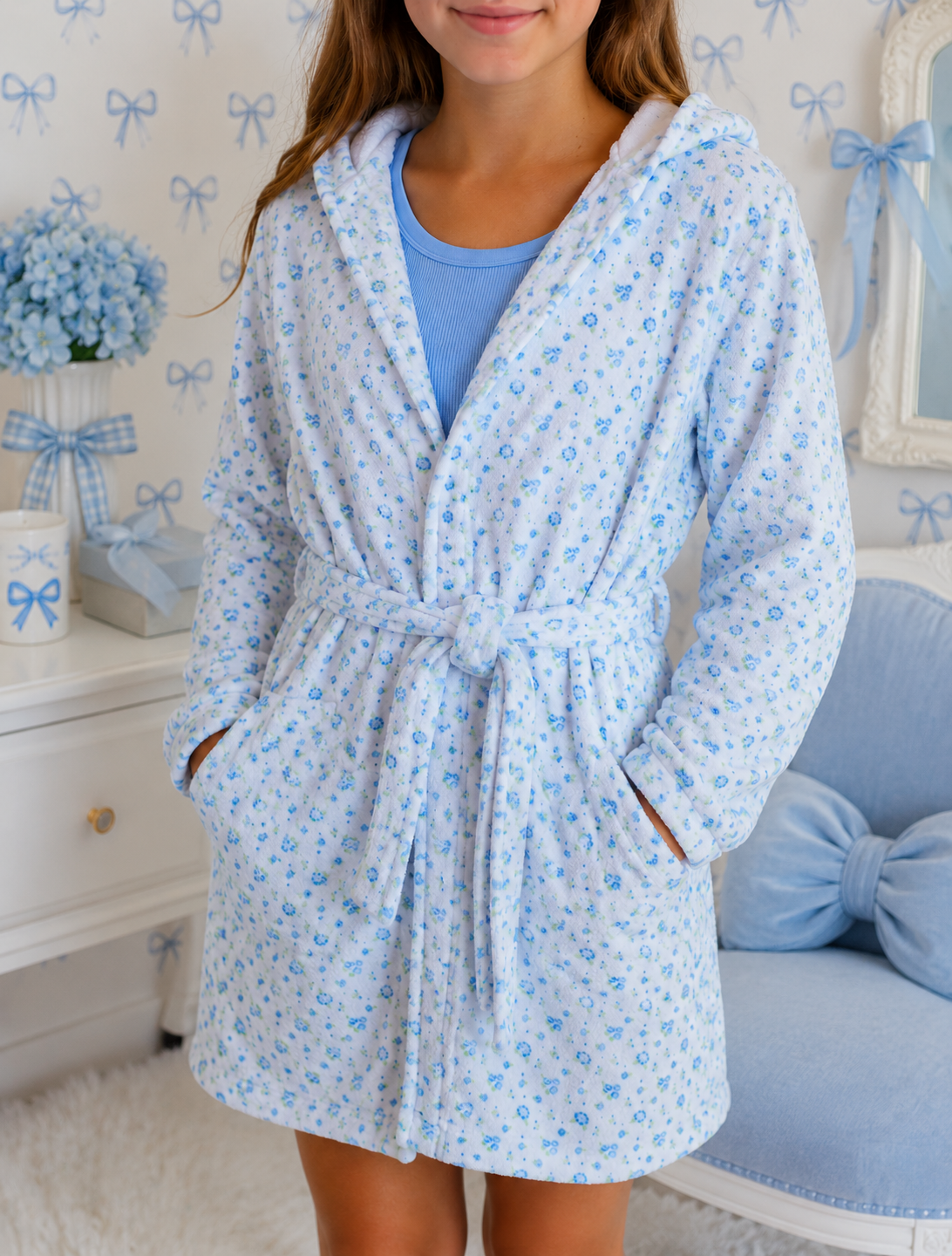 A young girl wearing an Iscream light blue floral print hooded robe over a light blue t-shirt, standing in a decorated bedroom setting with a blue chair and bow-patterned wallpaper.