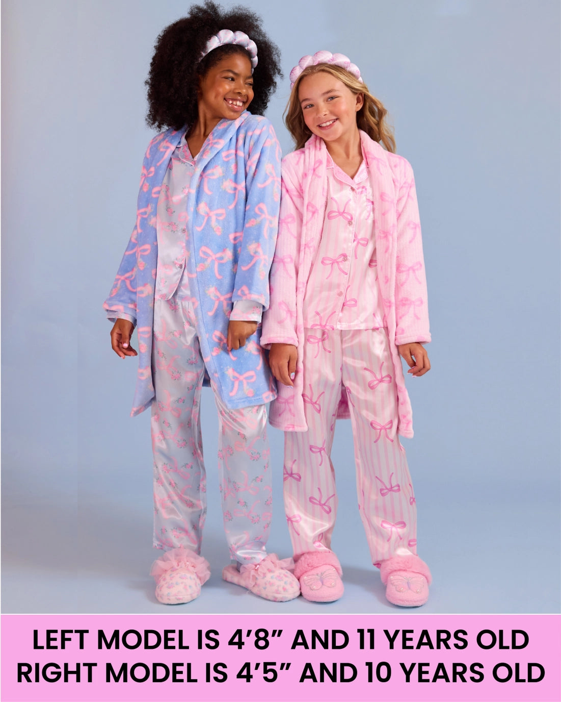 Two girls modeling matching pajama sets against a light blue-gray studio background. The left model wears a light blue robe and pants set with pink bow patterns and pink slippers, and the right model wears a pink robe and pants set with pink bow patterns and pink slippers.