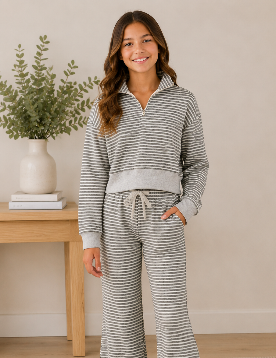 A young girl stands barefoot in a bedroom setting wearing a matching gray and white striped two-piece loungewear set consisting of a half-zip long-sleeve top and matching flared pants.