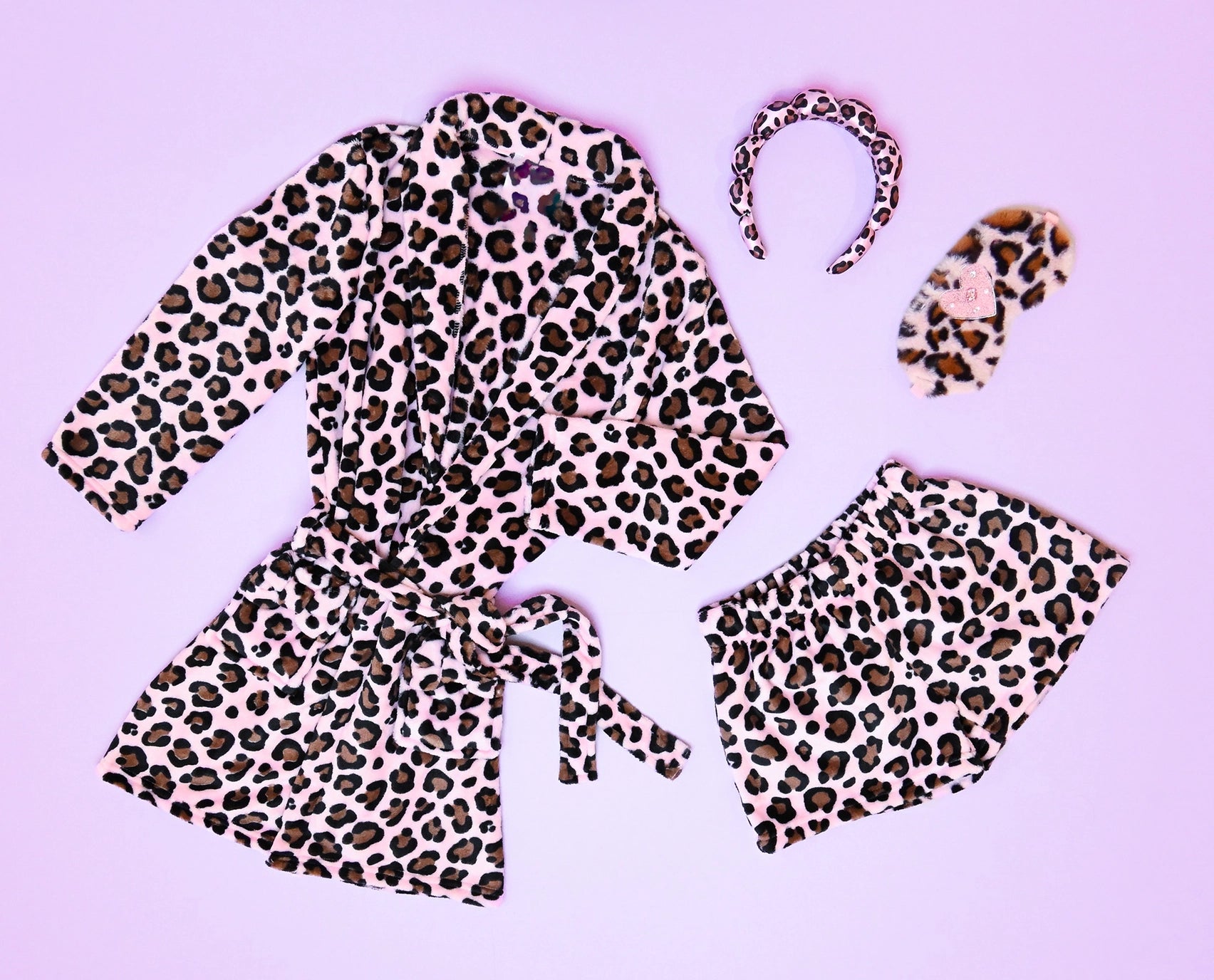 A matching three-piece gift set for girls that inlcludesloungewear set in a pink and brown leopard print, laid flat on a light purple background. The set includes a long-sleeve tie-front robe, a pair of shorts, and a headband.