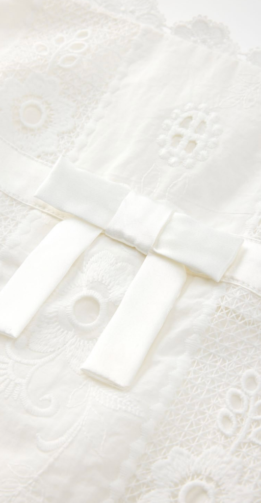 Close-up of a white embroidered lace garment with a satin bow detail, showing delicate eyelet and floral stitching.