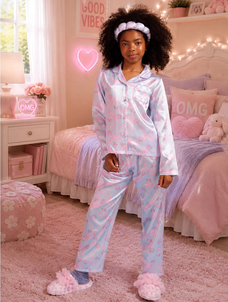 A young girl with curly hair stands in a pink bedroom wearing a light blue and pink patterned pajama set with a matching headband and fluffy pink slippers.