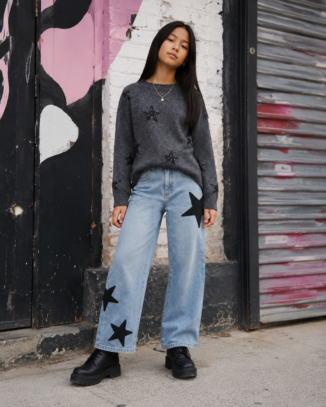 A girl stands outdoors in front of a graffiti-covered wall and metal shutter, wearing a dark gray knit sweater with black star patches by flowers by zoe, light blue wide-leg jeans with black star patches by KatieJ NYC , and black boots.