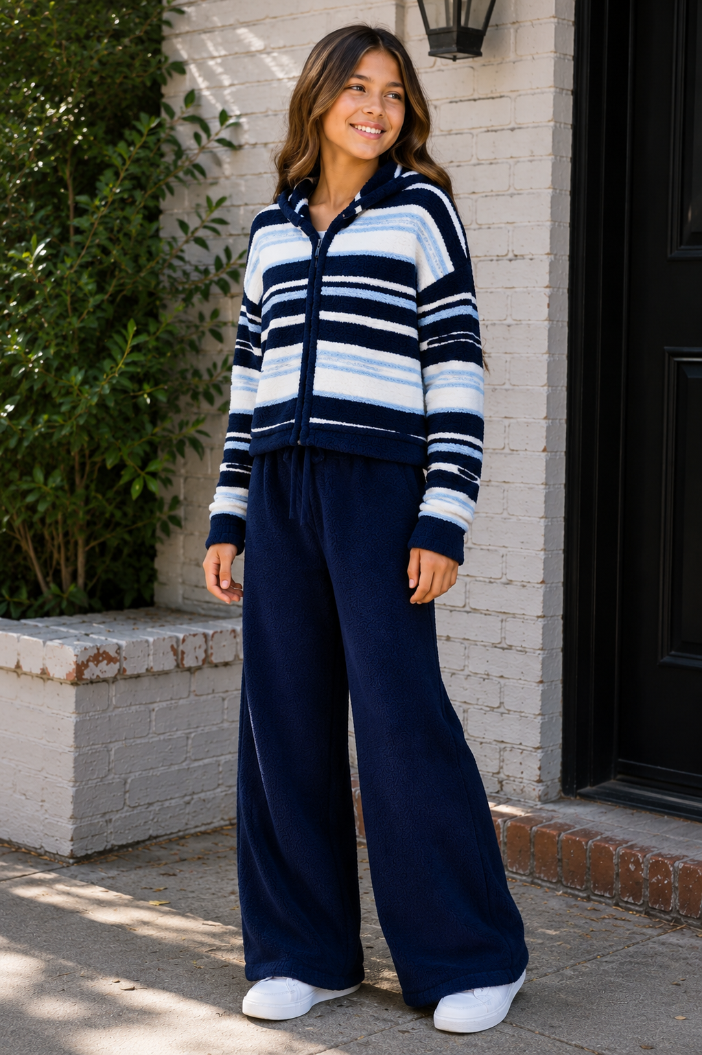 An 11 year old girl in a KatieJ outfits stands outdoors in front of a white brick wall and black door, wearing a navy, white, and light blue striped zip-up hoodie with matching navy wide-leg pants and white sneakers.