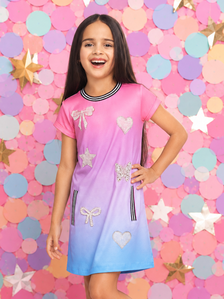 A young girl modeling a short-sleeve gradient dress that transitions from pink at the top to light blue at the bottom, decorated with white bows, hearts, and stars. She is standing in front of a colorful backdrop of pink, blue, and yellow confetti circles and gold stars.