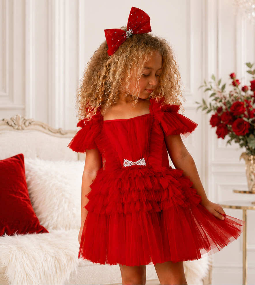 A young girl with curly blonde hair wearing a festive red off-the-shoulder tulle dress with a matching red bow in her hair, styled with white socks and black boots in an elegant room.