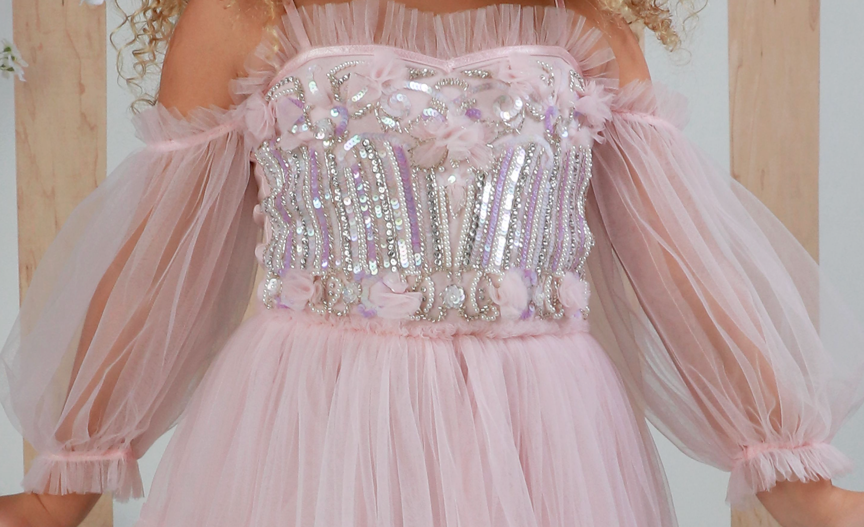 A close-up of a light pink tulle dress by Ooh! La, La! Couture with a heavily embellished bodice featuring silver sequins, rhinestones, and pink floral details, with sheer puff sleeves.