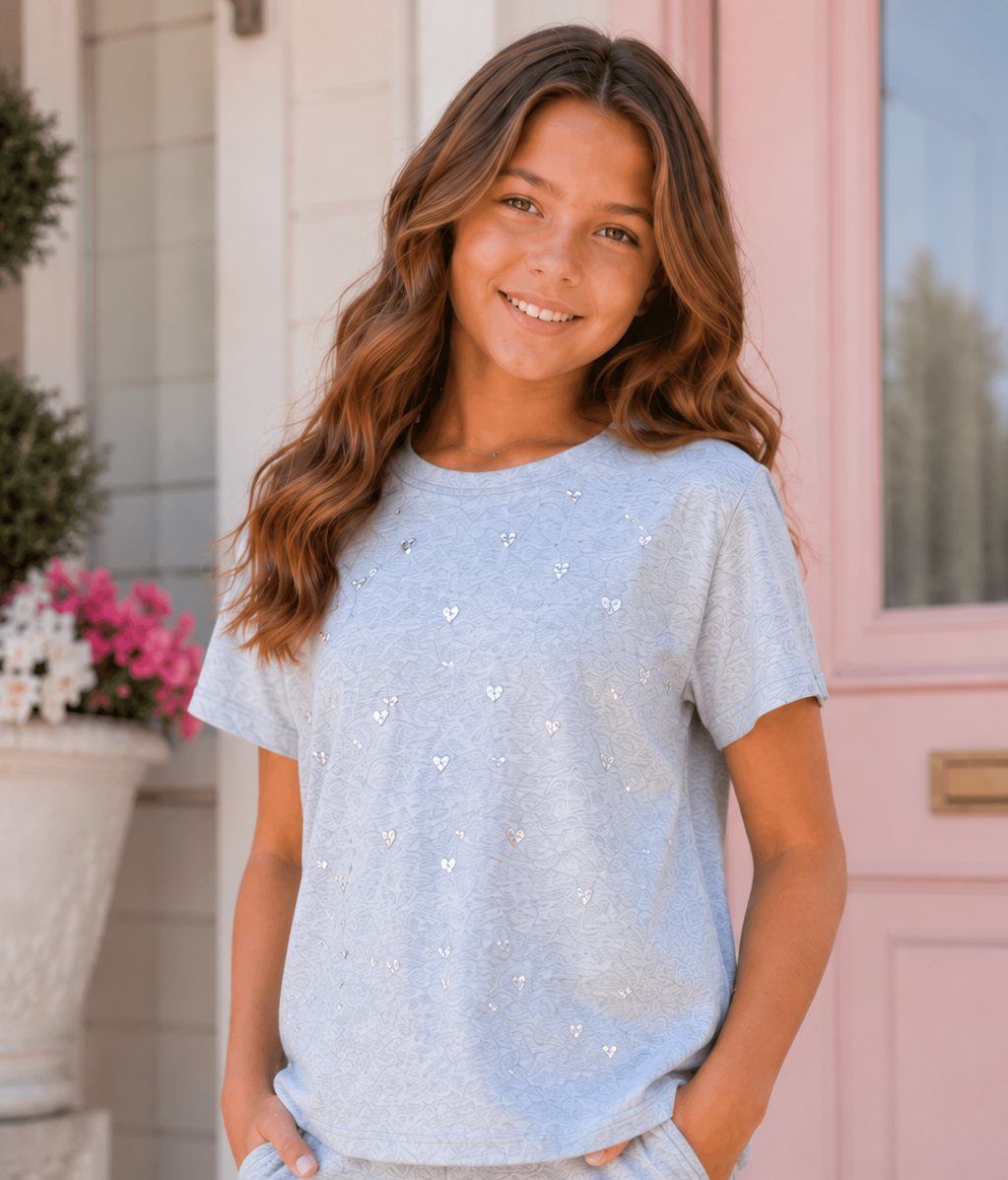 A young girl with long wavy brown hair stands smiling in front of a pink door, wearing a light heather blue short-sleeve t-shirt with small silver heart embellishments and matching heather blue sweatpants.