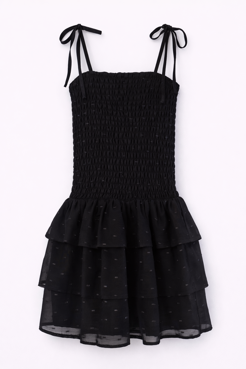 A black smocked girls mini dress with thin tie-shoulder straps, a smocked bodice, and a tiered ruffled skirt with a subtle polka dot pattern, shown against a plain white background.