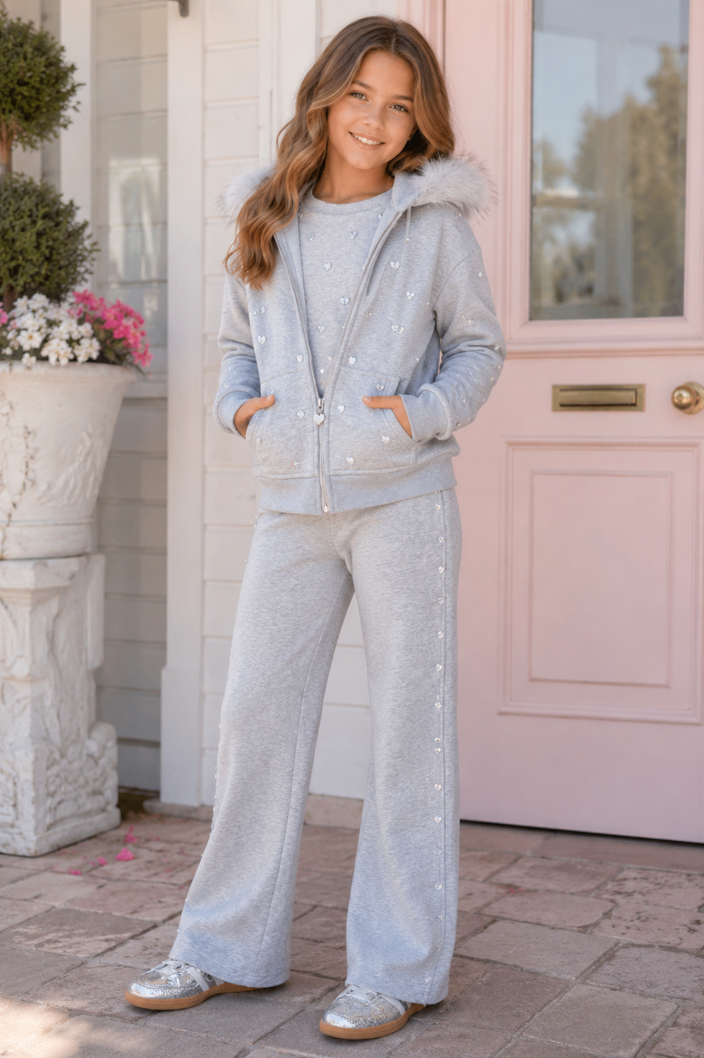 A young girl stands on a front porch wearing a light gray heathered two-piece loungewear set with silver rhinestone embellishments. The set includes a zip-up hoodie with a faux-fur trimmed hood and matching wide-leg sweatpants.
