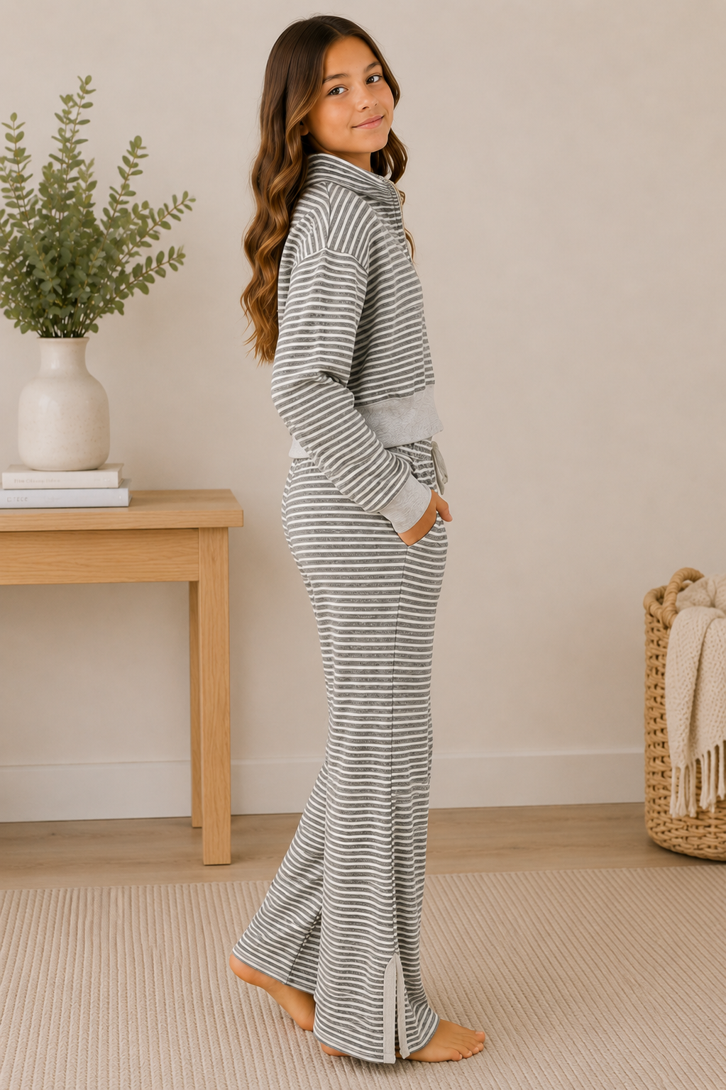 A young girl modeling a matching gray and white striped lounge set, consisting of a long-sleeve turtleneck top and matching flared pants, standing in a brightly lit room with a wooden side table and a plant in the background.