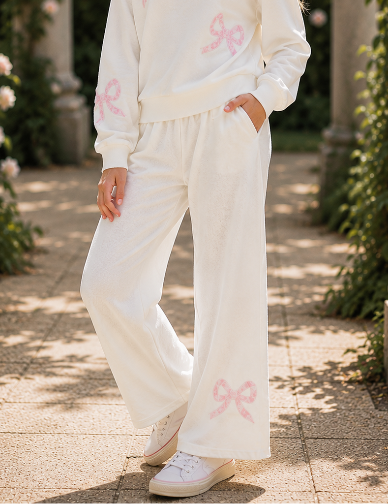 A young girl stands in a sunlit garden walkway wearing a matching white lounge set with pink bow prints. The set includes a long-sleeve sweatshirt and wide-leg pants, styled with white sneakers.